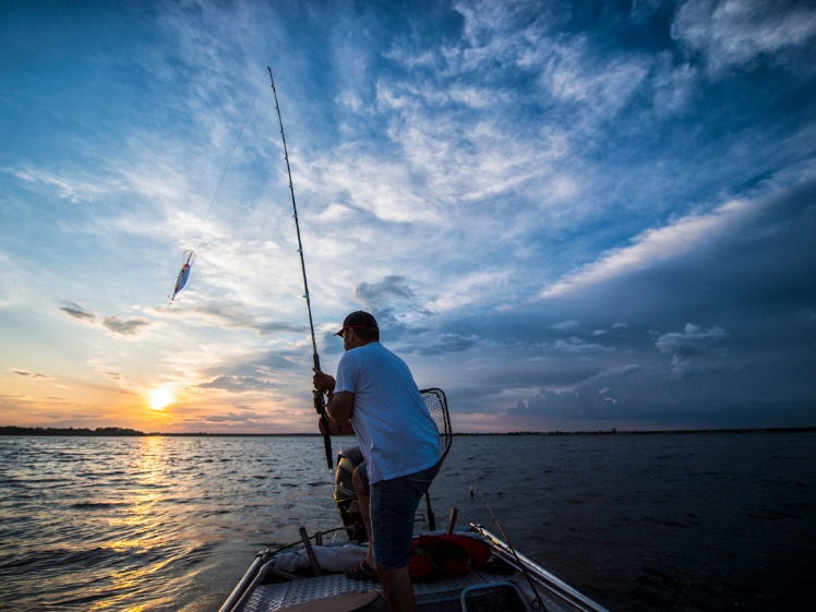 A man on his boat, casting his fishing rod during sunset.